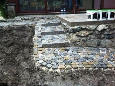 Teilweise gepflasterter Gartenweg mit Natursteinen, umgeben von aufgelockerter Erde.