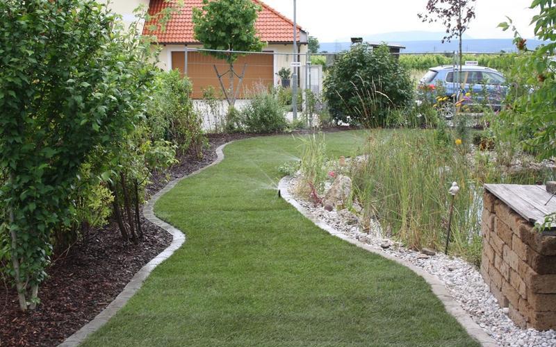 Geschwungener Rasenweg mit Beetbegrenzungen aus Steinen, gesäumt von Sträuchern.