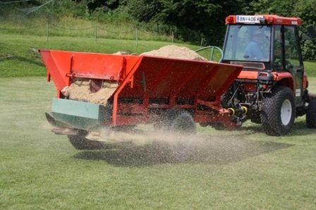 Traktor mit Sandstreuer verteilt Bodenmaterial auf einer Rasenfläche.