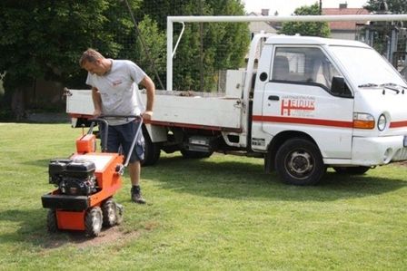 Mitarbeiter mit einer kleinen Bodenfräse bearbeitet eine Rasenfläche neben einem weißen Transporter.
