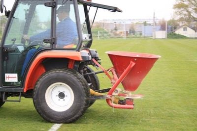 Traktor mit einem Streuwagen verteilt Dünger oder Sand auf einem Sportfeld.